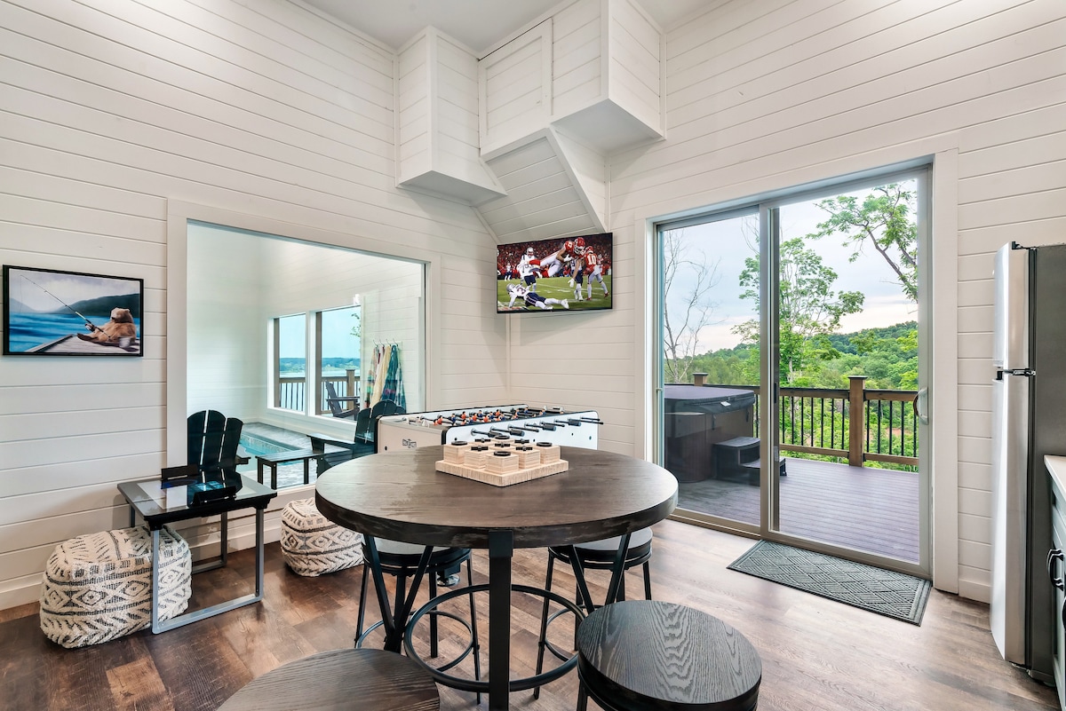 An open dining area features a round table with black seating for six. A large mirror reflects the natural light from the sliding glass doors, which lead to a deck. A foosball table is visible, along with a television displaying a sports game.