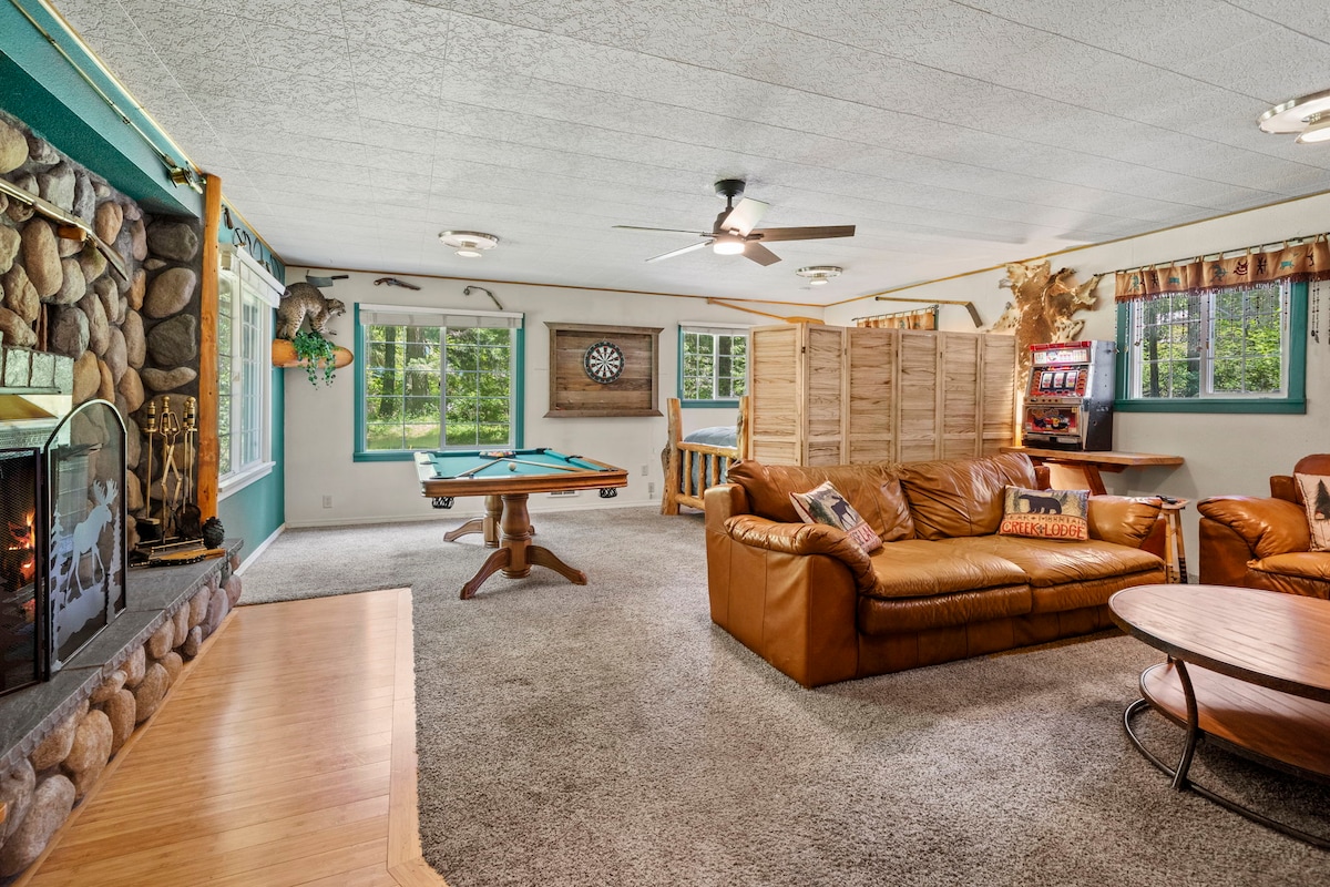 A spacious indoor living area is enhanced by large windows that invite natural light. Comfortably arranged leather couches surround a central coffee table, while a pool table and dartboard provide recreational options. A stone fireplace adds warmth, complemented by a retro slot machine for entertainment.