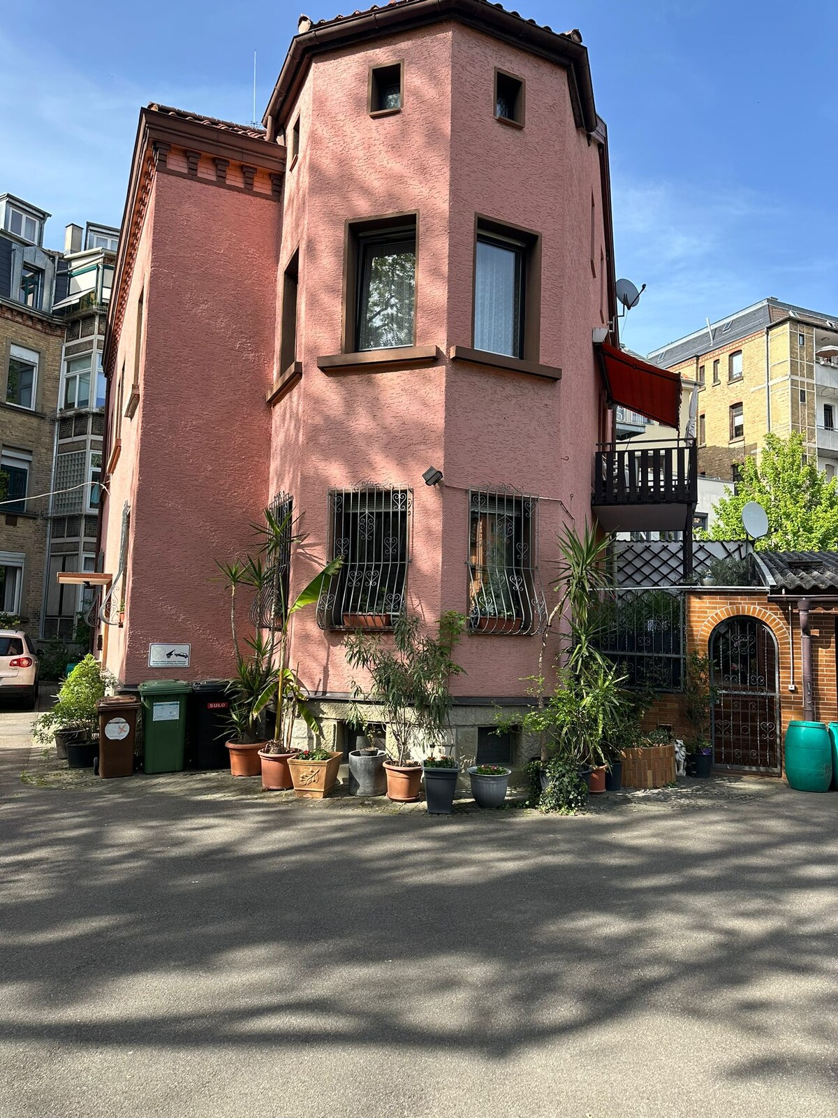 A charming pink building features a distinctive turret and multiple windows, framed by greenery and potted plants. The ground level presents an organized space with bins and a stone pathway, inviting guests into a welcoming environment.