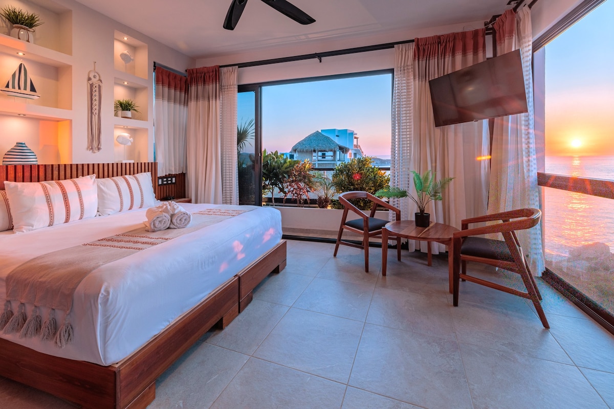 A spacious room features large windows with breathtaking ocean views at sunset. A King size bed is accompanied by two rolled towels. A small table with two chairs sits by the window, while light drapes enhance the serene atmosphere.