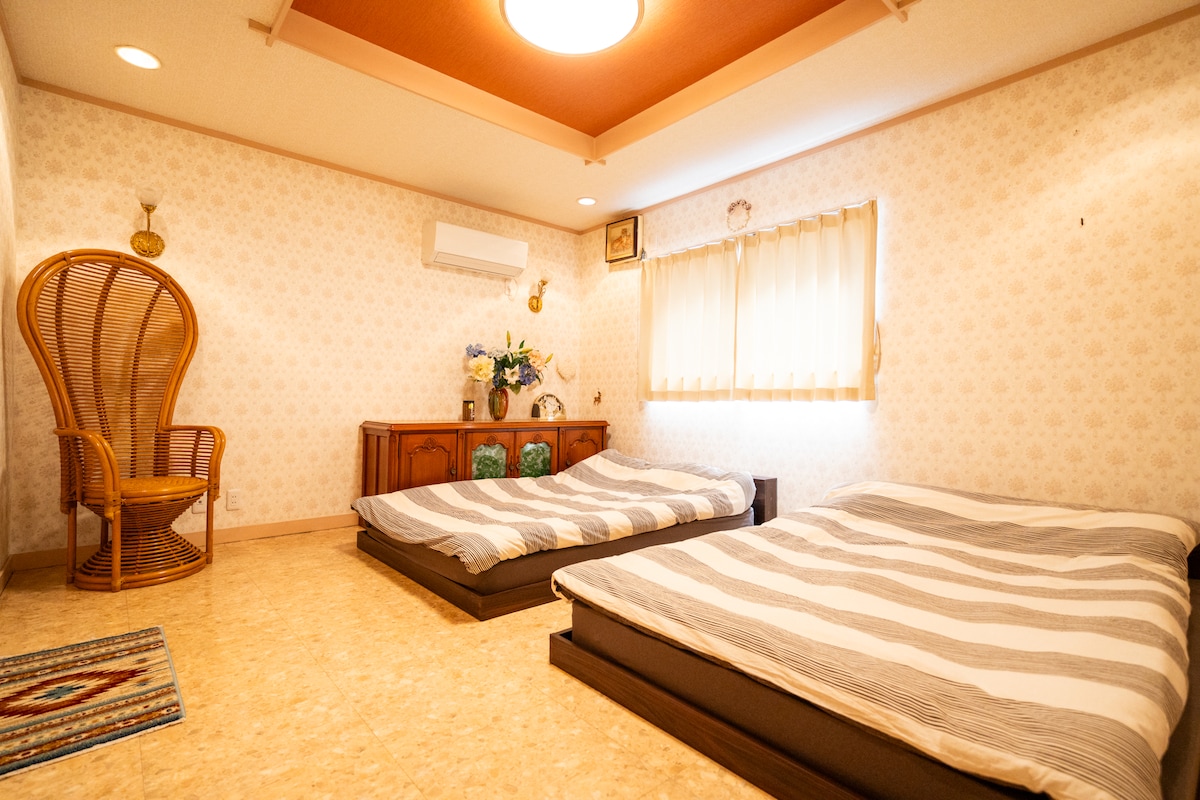 The room features two spacious beds with striped linens positioned on a smooth surface. A decorative chair stands in one corner, while a wooden cabinet with floral arrangements adds warmth. Natural light filters through window treatments, creating a calming ambiance.