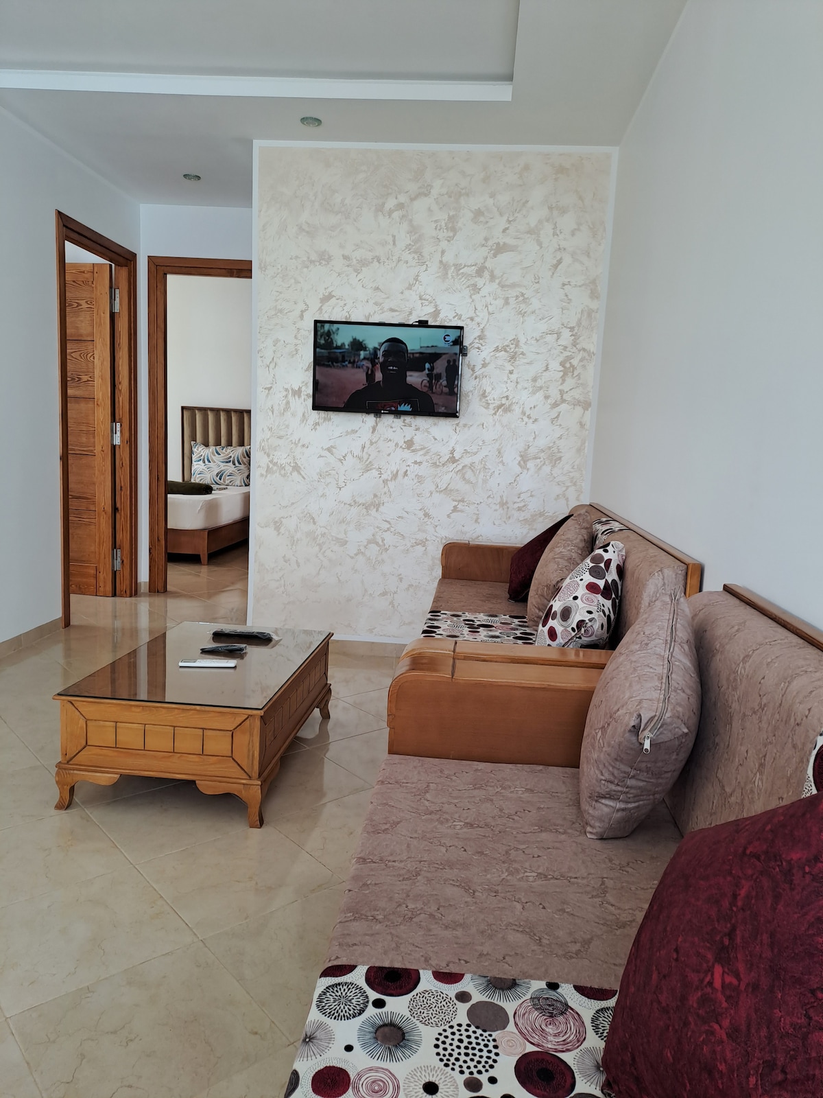 A comfortable living area is seen with a brown sofa and patterned cushions, complemented by a glass coffee table. A wall-mounted television is visible, and a doorway leads to a bedroom, partially obscured. The tiled floor reflects natural light, enhancing the room's openness.