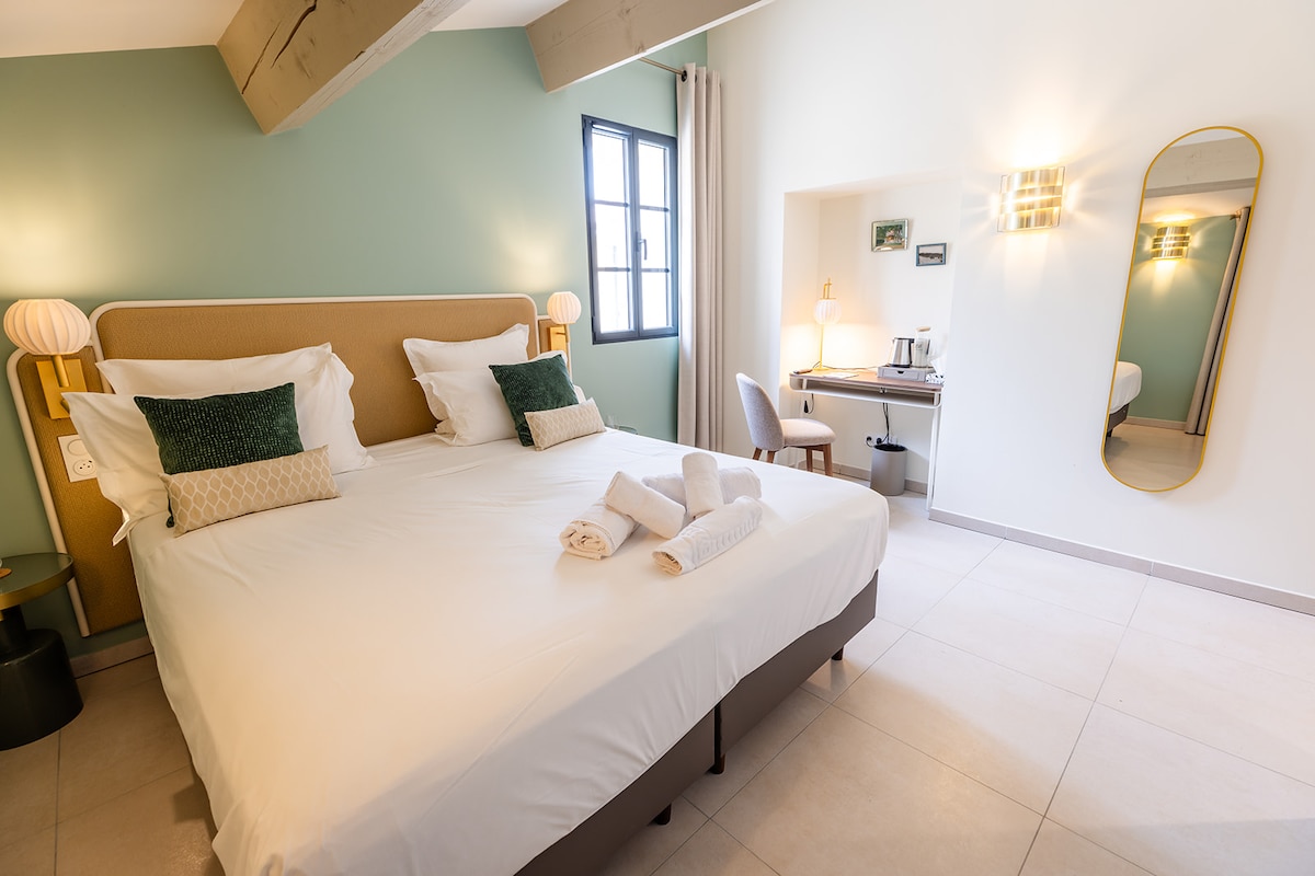 A spacious room is featured with a large bed adorned with decorative pillows. A work desk with a chair is positioned against the wall, accompanied by a kettle and accessories. Natural light enters through a window, while soft lighting highlights the room's pastel colors.