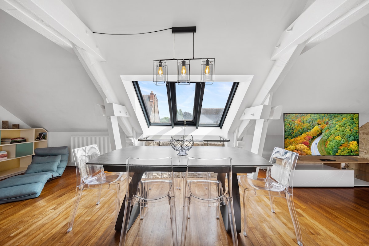 A spacious dining area features a large table surrounded by transparent chairs, enhancing the open feel. Large skylight windows fill the room with natural light, showcasing wooden beams and modern decor. A cozy seating option is positioned nearby, complemented by a wall-mounted television.