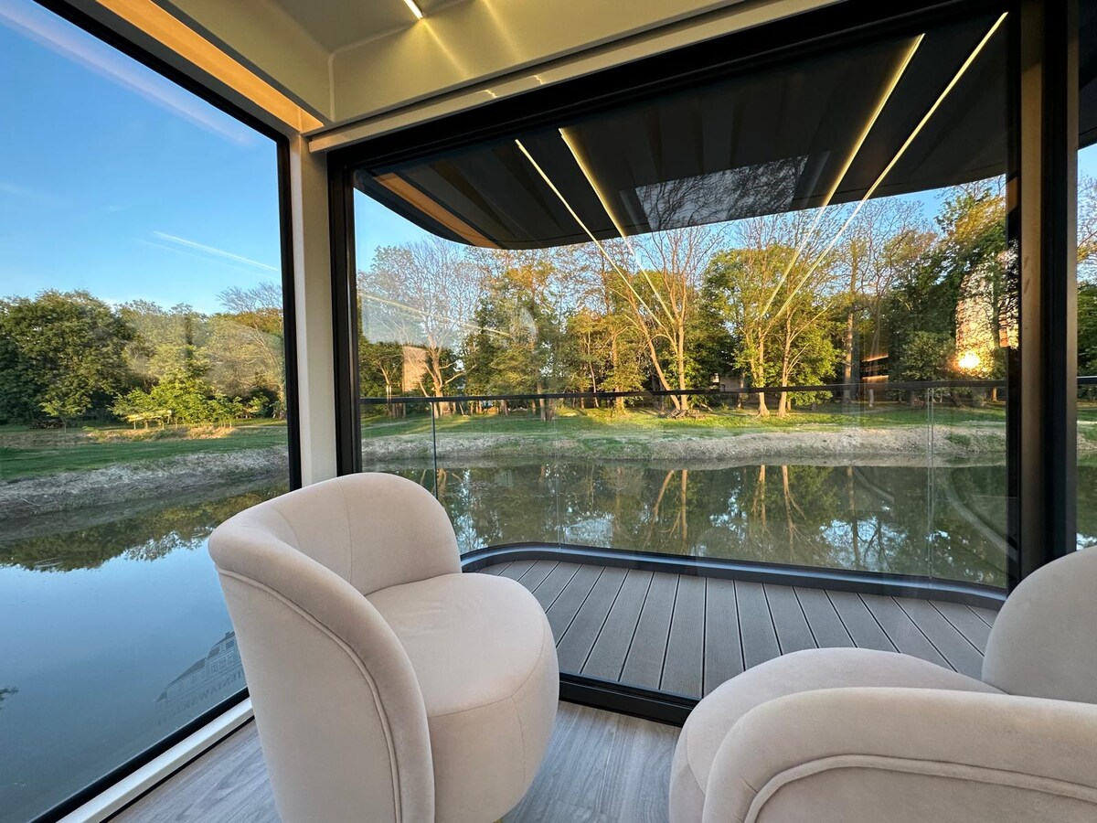 Two plush armchairs are positioned on a wooden deck, offering an unobstructed view of a serene body of water surrounded by trees. Large glass windows allow natural light to flood the space, creating a calming atmosphere.