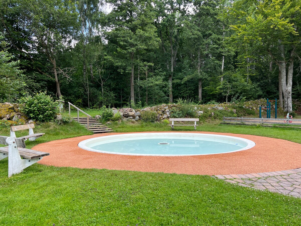 A circular outdoor pool is surrounded by a green lawn and trees, providing a peaceful setting. Benches are positioned nearby for relaxation. Access to the pool is facilitated by a set of steps leading through the grassy area.