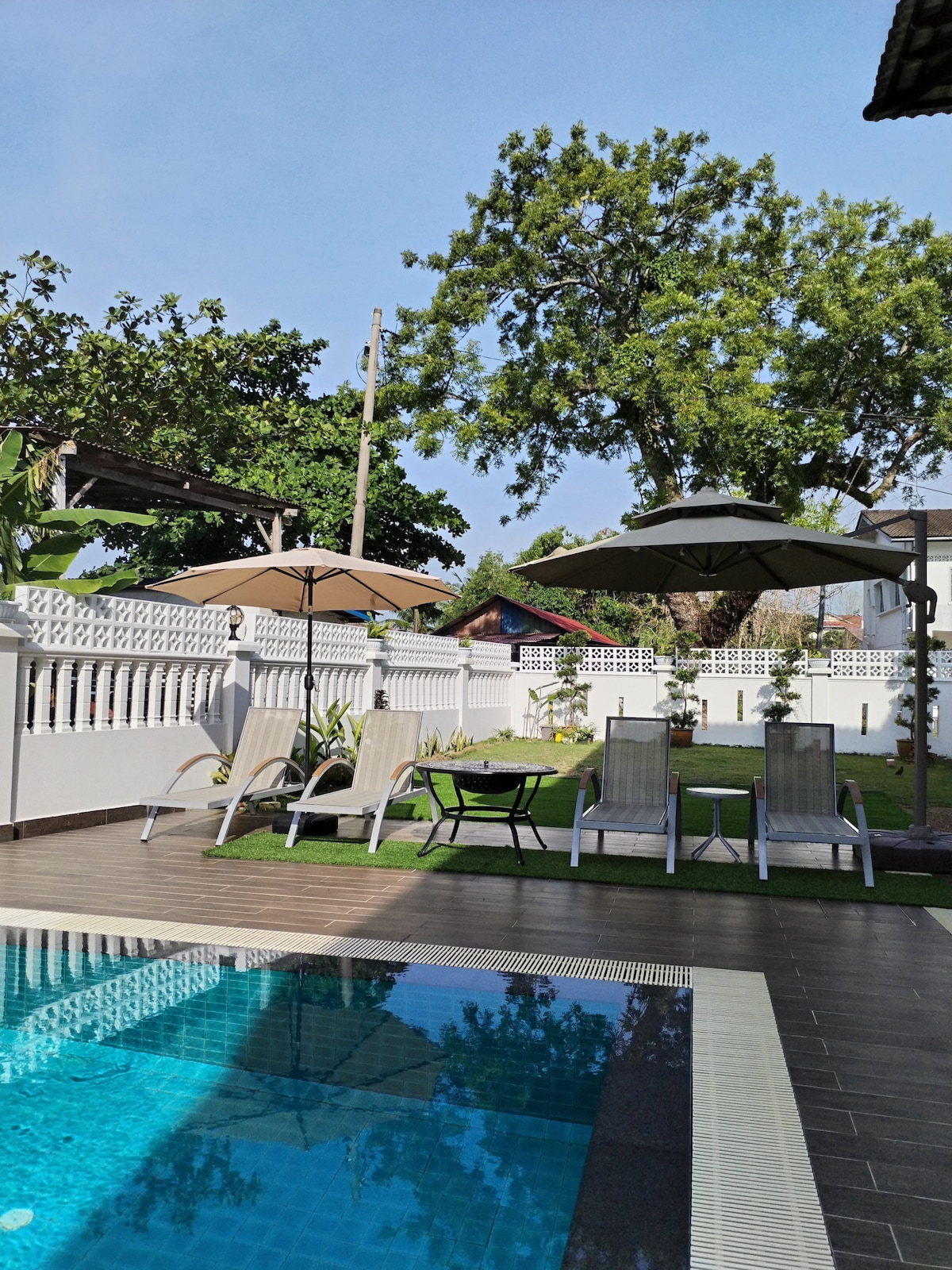 A private pool area is shown, with crystal-clear water reflecting the surroundings. Two lounge chairs are positioned beside the pool, along with a small dining table shaded by an umbrella. Lush greenery and plants line the perimeter, enhancing the outdoor space.