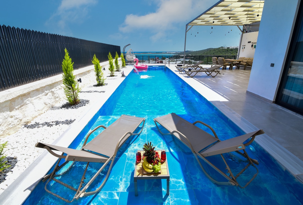 A sunlit pool area features two reclining lounge chairs positioned beside a clear blue pool. A decorative table holds tropical fruits and drinks, while greenery is planted along the edges. A spacious outdoor dining area is visible under a shaded pergola in the background.
