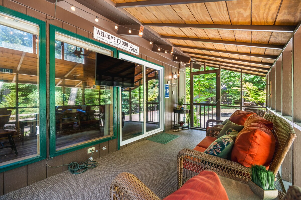 A screened-in porch features comfortable seating with vibrant orange cushions. A wall-mounted television is visible, and greenery can be seen through large windows, creating a bright and inviting space. Warm lighting from string lights above adds to the welcoming ambiance.
