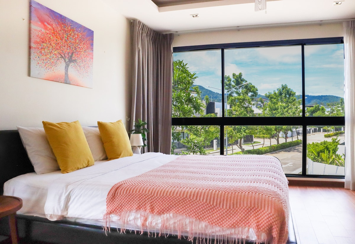 A serene bedroom features a spacious bed adorned with soft pillows and a textured blanket draped at the foot. Large windows provide ample natural light and showcase views of lush greenery and a pathway. A calming wall art piece adds a touch of color.