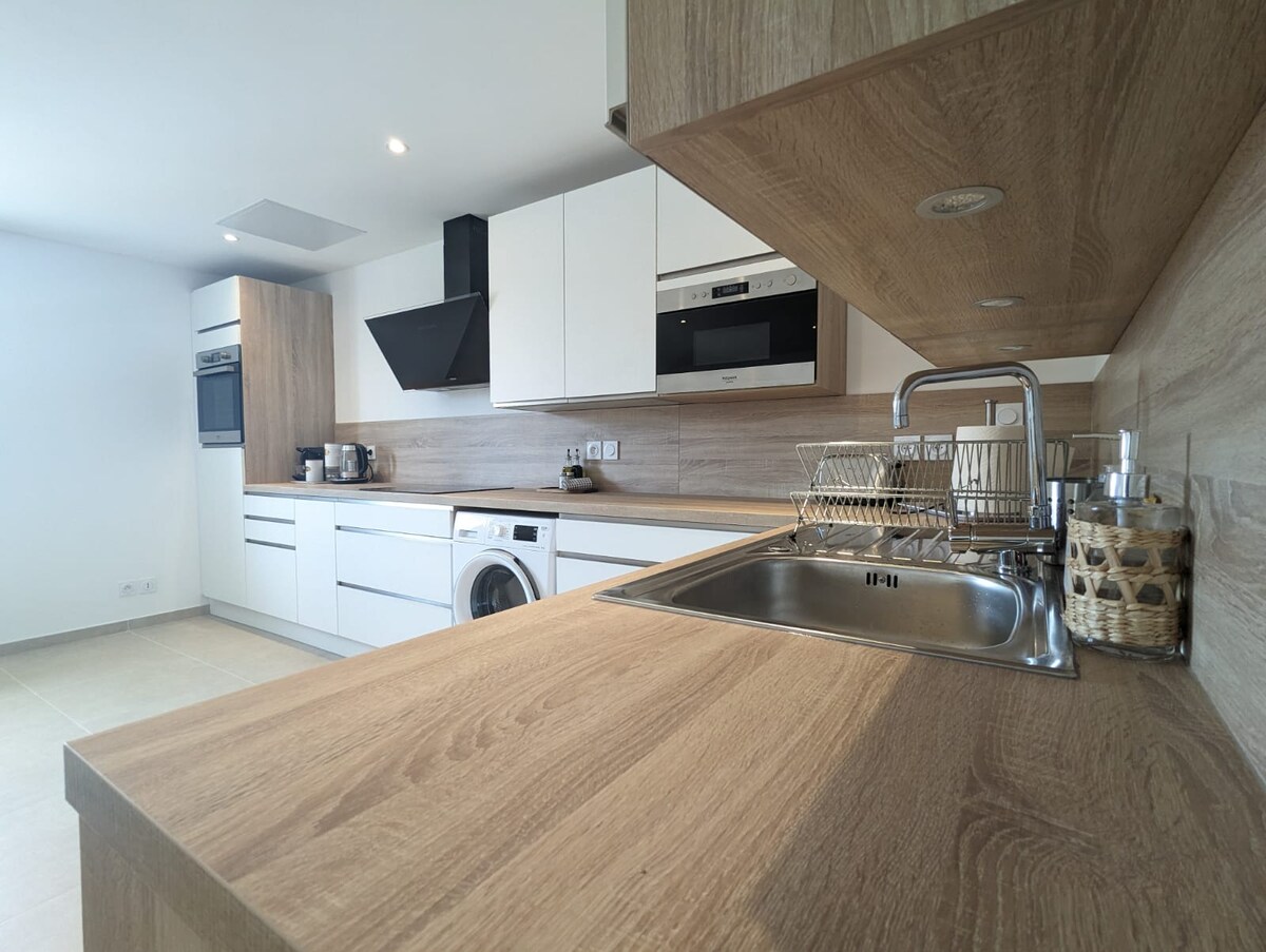 The kitchen features a sleek design with light wood cabinetry and modern appliances. A stainless steel sink is positioned next to a drying rack, and a washing machine is integrated within the cabinetry. Ample counter space is available for meal preparation.