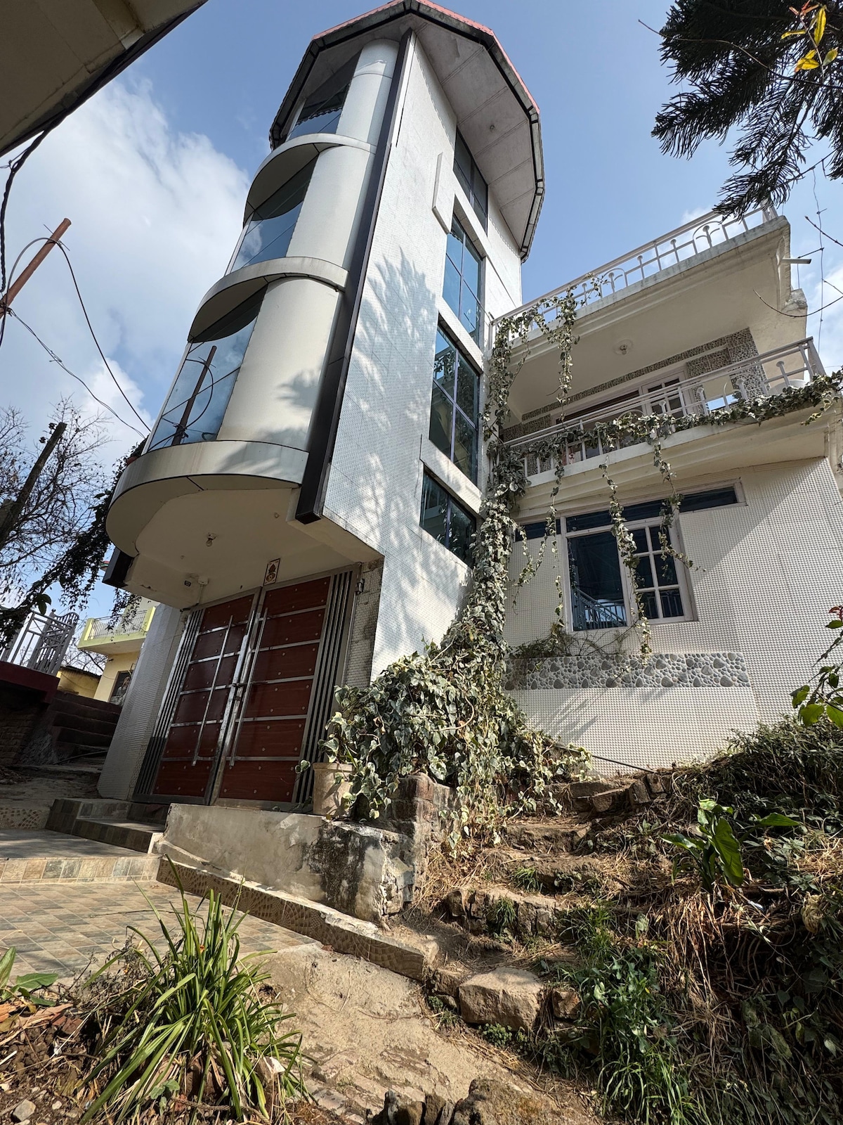 A multi-level villa is shown, featuring a modern design with large windows. The exterior is accented by creeping vines, and a pathway leads to the entrance. The structure stands amidst a natural setting, complemented by greenery and trees.