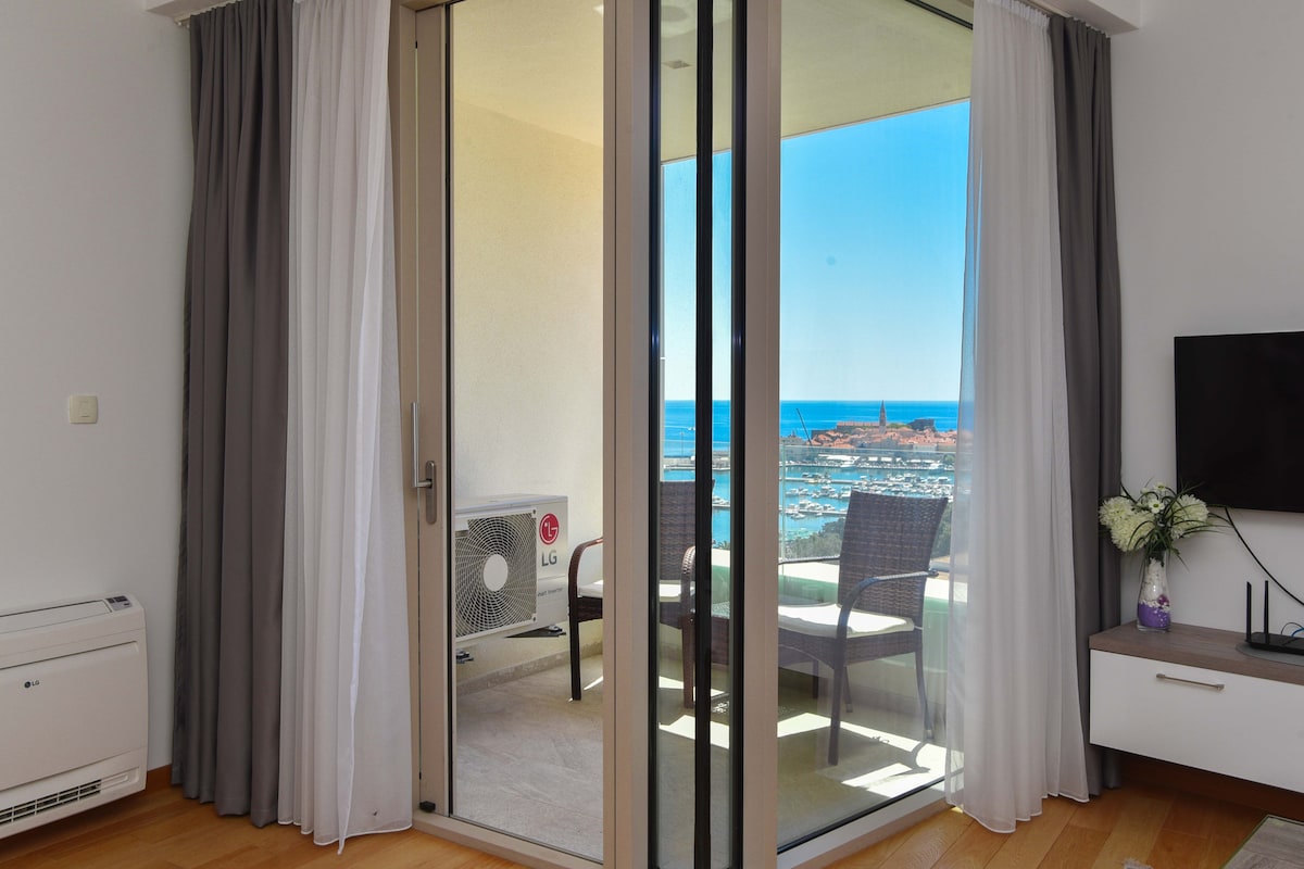 A glass door opens to a private balcony with a view of the sea. Light curtains frame the entrance, and outdoor seating is visible. A TV and a small table are located in the interior space, complemented by light-coloured wooden flooring.