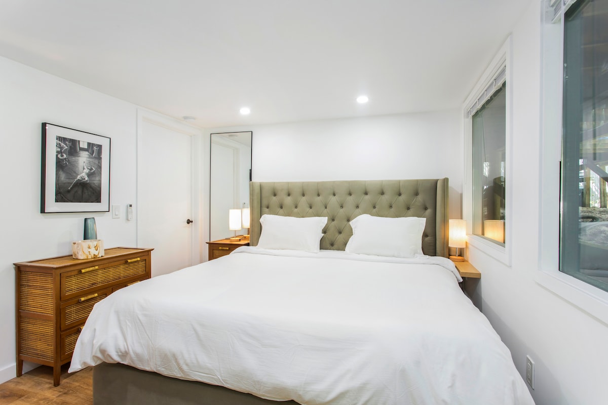 A well-appointed sleeping area features a king size bed with light bedding and soft pillows. Two bedside lamps provide warm illumination. A framed black-and-white photograph is hung on the wall, and a dresser adds functional storage against the bright white backdrop.