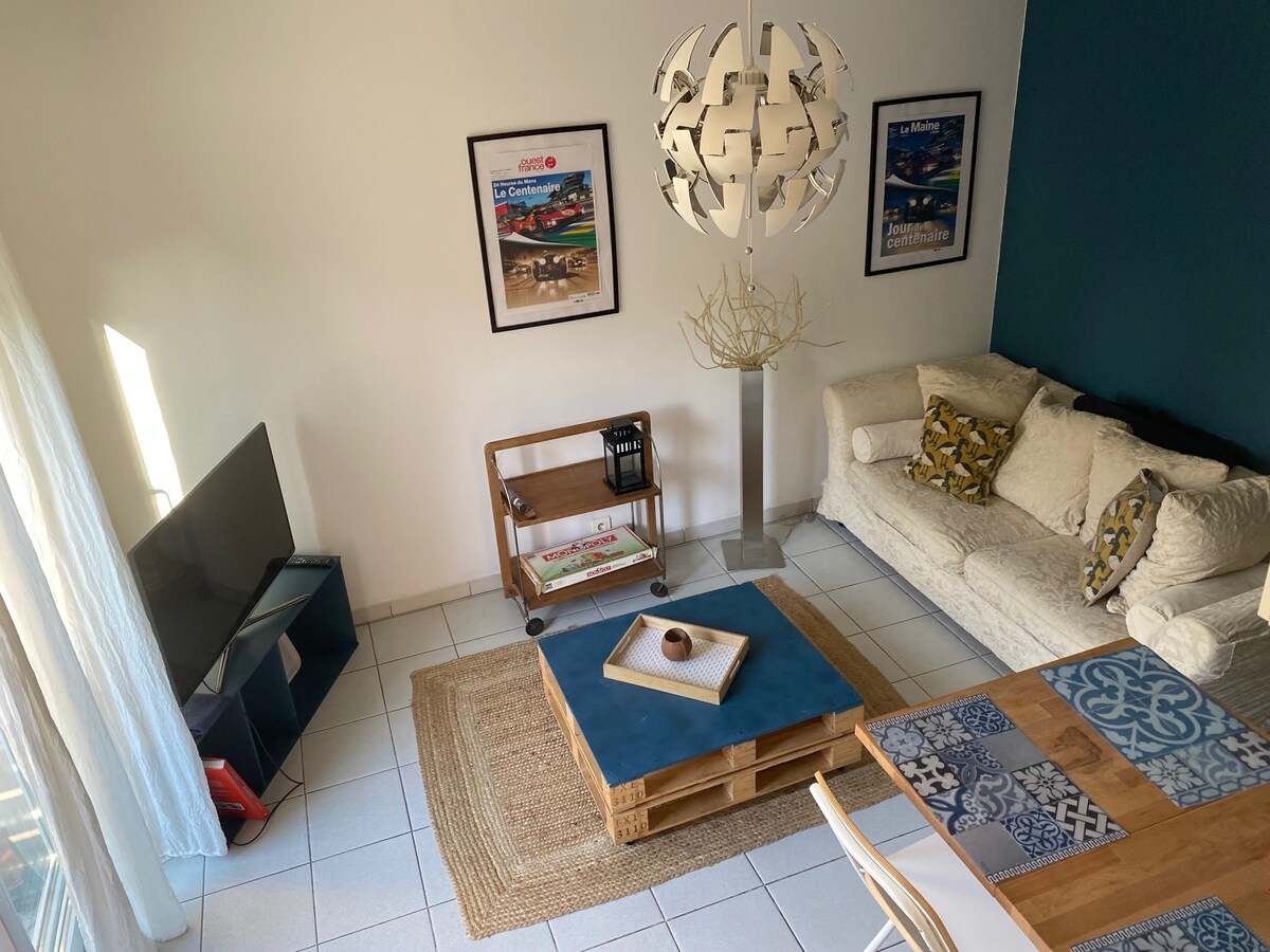 A light-filled living area features a comfortable white sofa adorned with patterned cushions. A wooden coffee table occupies the center, complemented by a decorative bowl. On one side, a sleek TV stands on a modern cart, while artistic prints decorate the walls, enhancing the room's character.