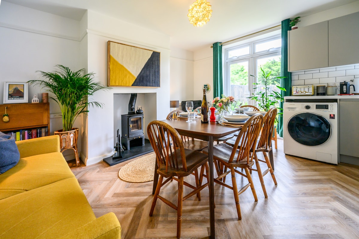 An open-plan living and dining area is showcased, featuring a wooden dining table set for meals, surrounded by six chairs. A cozy wood-burning stove adds warmth to the space, while large windows allow natural light to fill the room. The modern kitchen is visible in the background.