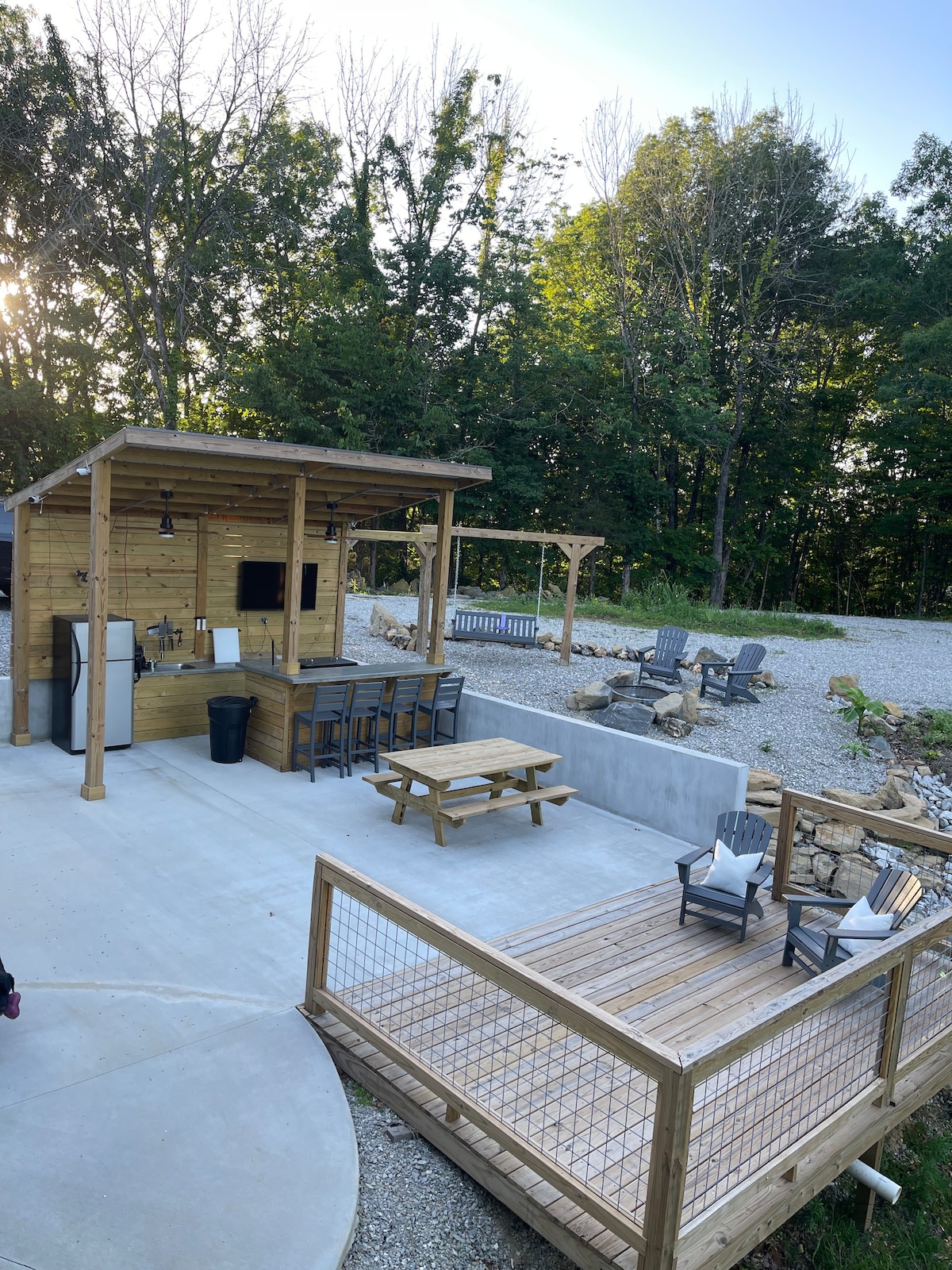 An outdoor kitchen area is featured with a wood structure including a grill and bar seating. Several lounge chairs are arranged around a spacious concrete area, complemented by natural stone landscaping and greenery in the background. A small elevated deck offers additional seating options.
