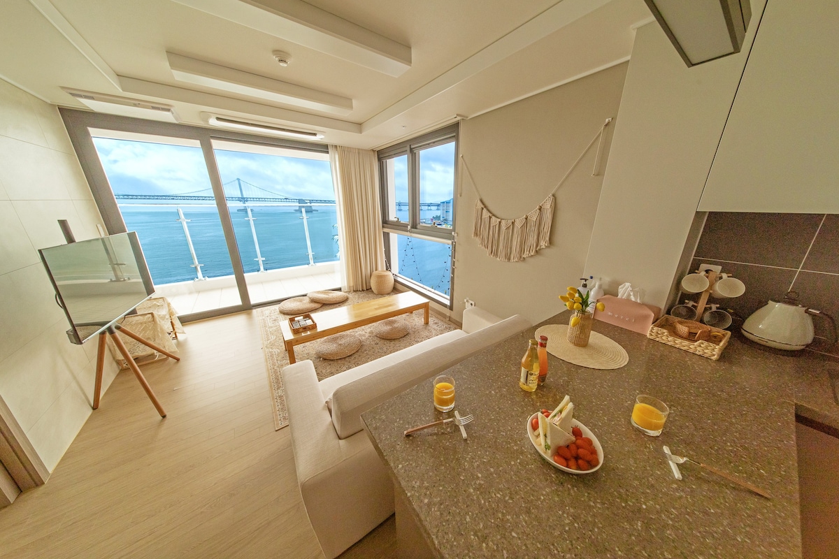 A bright living area is showcased, featuring a spacious sofa and a stylish kitchen counter. Large windows offer stunning views of the ocean and Gwangandaegyo Bridge. A small table is set with fruit and drinks, enhancing the inviting ambiance.