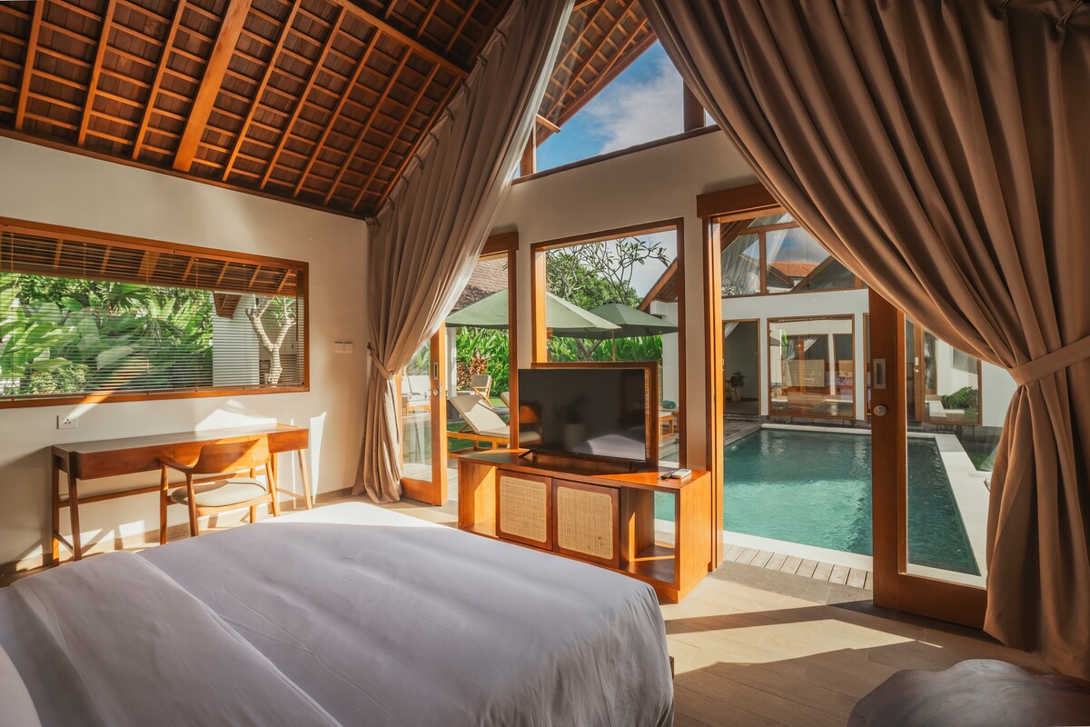 A bright bedroom features large windows with natural light flooding in. A spacious bed is positioned in the foreground, while a wooden desk and chair are visible. The view extends to a tranquil pool area surrounded by greenery, creating a serene atmosphere.