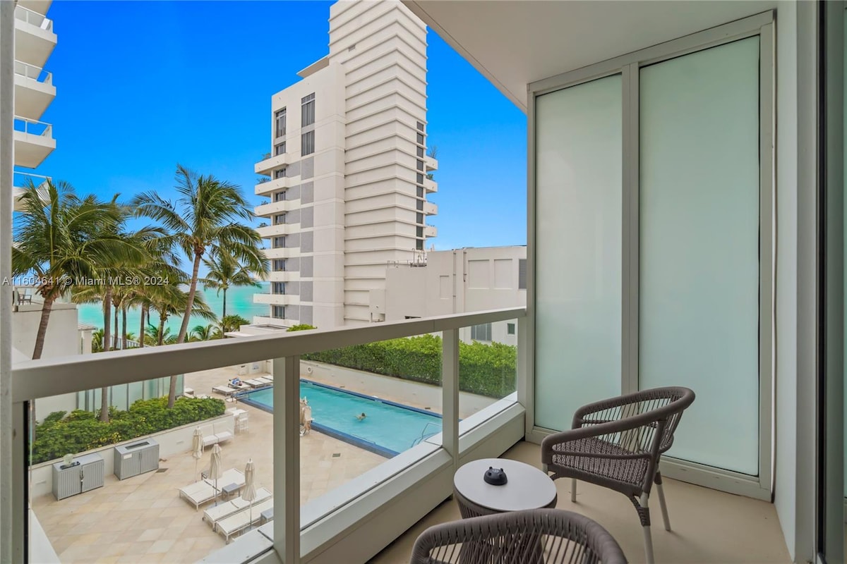 A private balcony features two stylish chairs overlooking a pool area surrounded by palm trees. The clear blue sky complements the contemporary architecture of the nearby buildings, creating a serene space for relaxation.