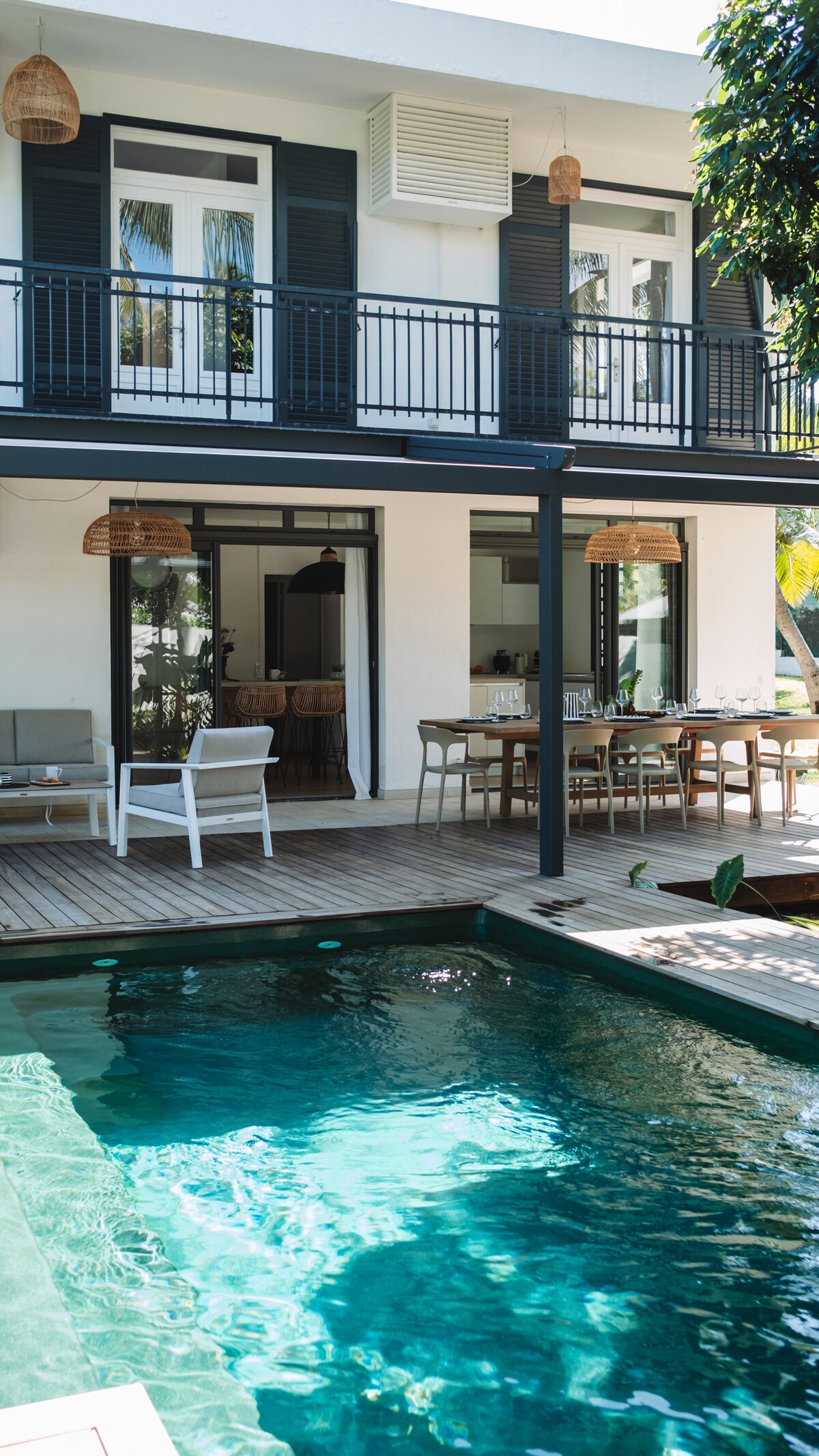 An outdoor area features a turquoise pool surrounded by wooden decking. Comfortable seating is provided under a shaded space, with a dining table visible in the background. Large windows frame the indoor space, enhancing the connection between inside and outside.