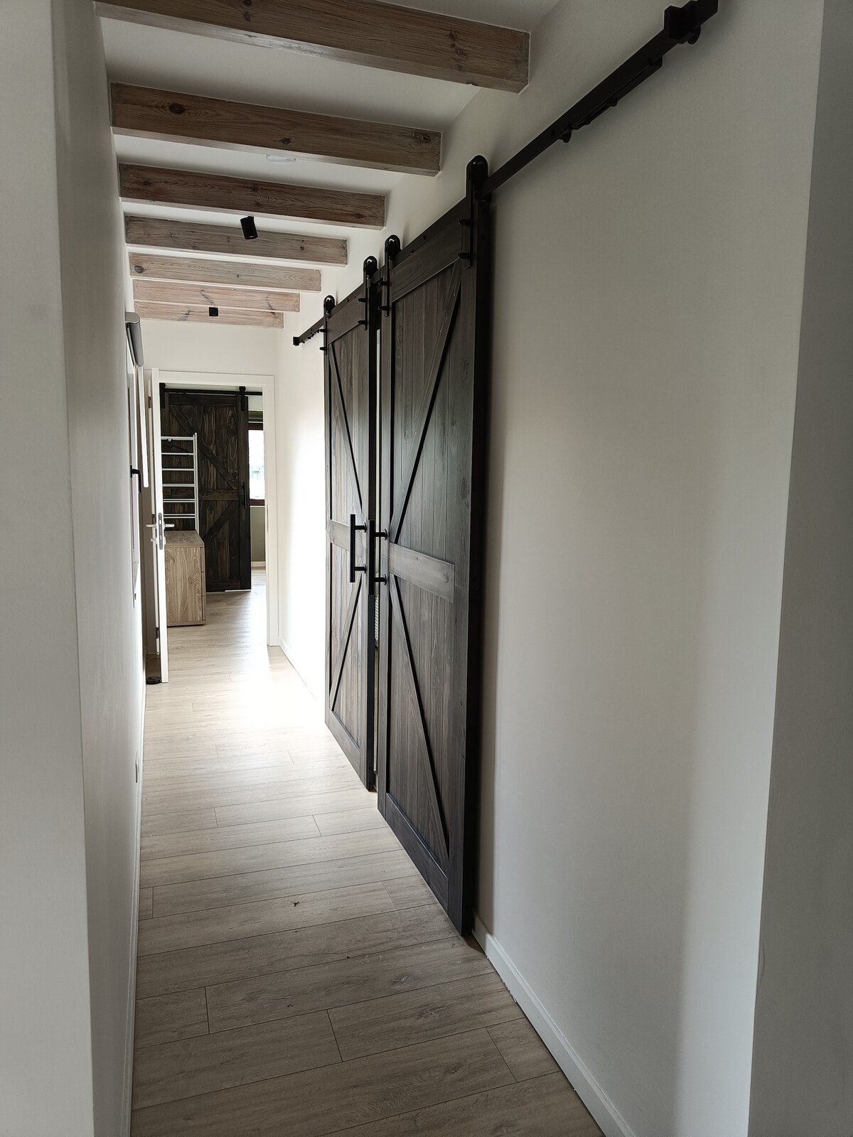 A light-filled hallway is presented, featuring a smooth wooden floor and exposed beams overhead. Sliding barn doors made of dark wood are positioned on one side, leading to other areas of the chalet. Soft lighting enhances the inviting ambiance throughout the corridor.