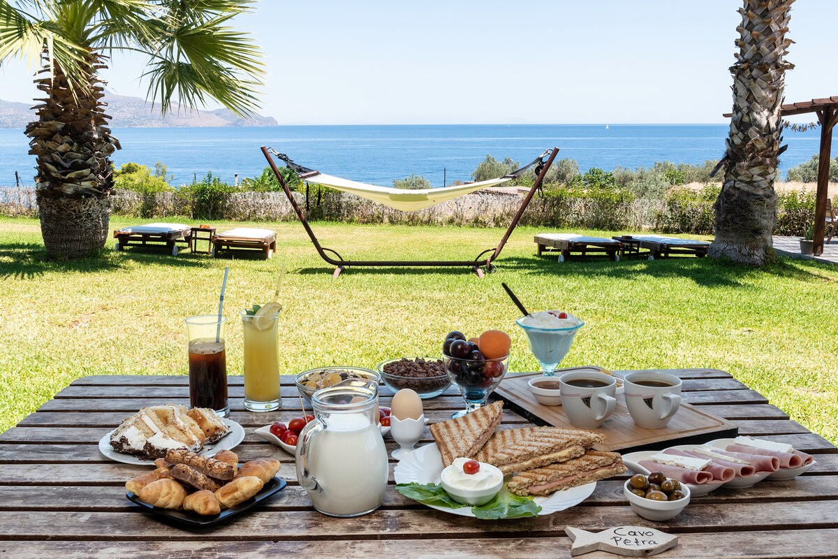 A large wooden table is set outdoors, displaying a variety of beverages and breakfast items. In the background, a hammock is suspended between two palm trees, with a clear view of the blue sea and distant shoreline. Lush greenery surrounds the area, enhancing the tranquil setting.