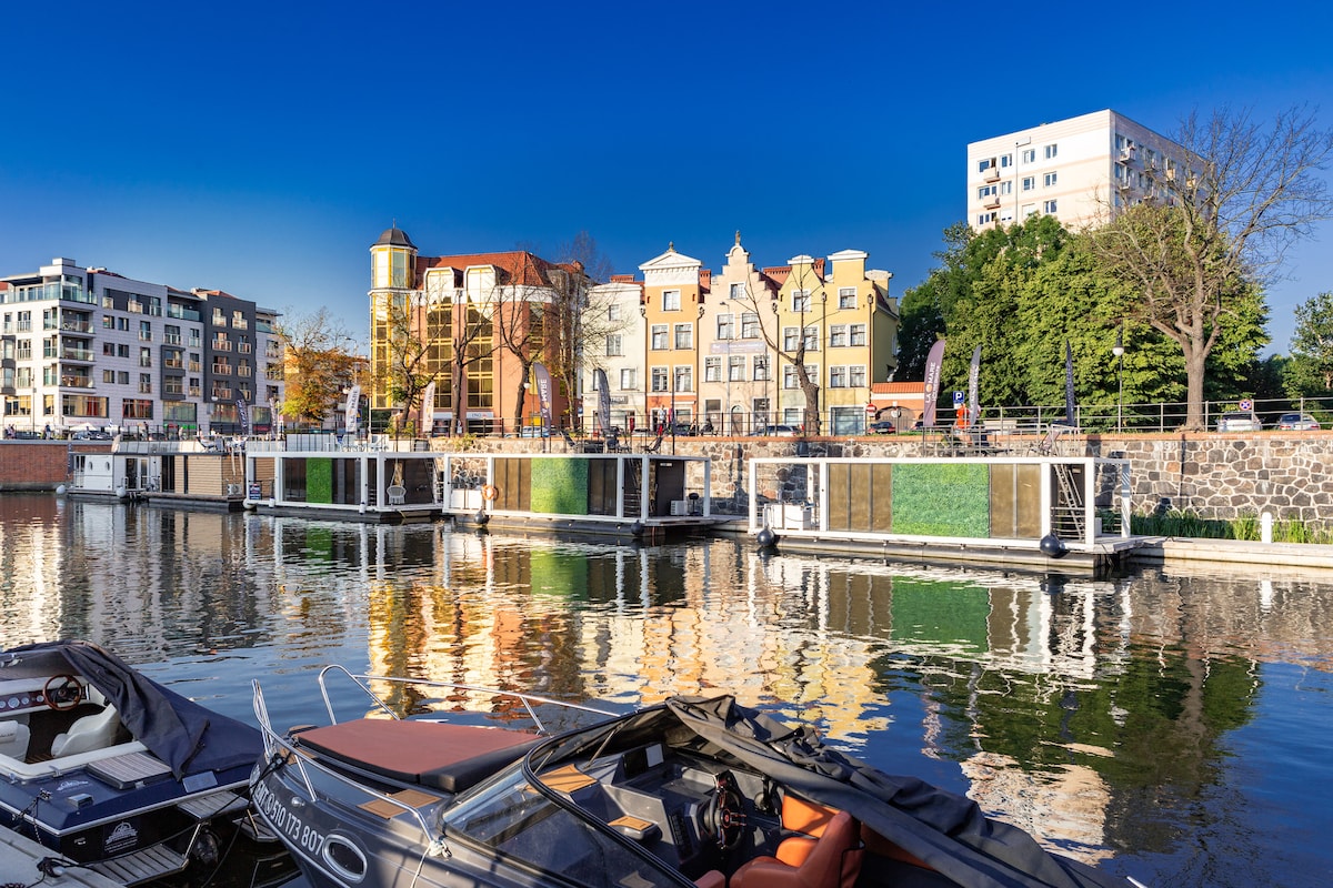 A serene waterfront scene features a line of houseboats moored along the dock, their colorful exteriors reflecting in the calm water. The backdrop includes a variety of buildings showcasing architectural diversity under a clear blue sky, creating a pleasant atmosphere.
