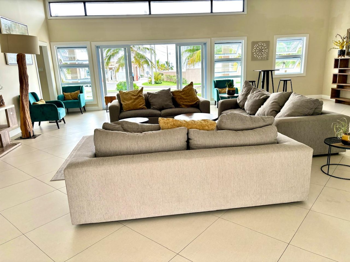 A spacious living area is furnished with comfortable seating, featuring a light grey sectional sofa and accent chairs in vibrant shades of teal. Large windows allow natural light to fill the space, providing views of palm trees and the outdoor environment.