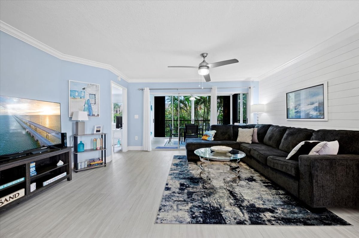 The living area features a spacious sectional sofa arranged around a glass coffee table. A large flat-screen TV is positioned beside a bookshelf. Natural light floods the room from the sliding glass doors, which open to a balcony overlooking lush greenery.
