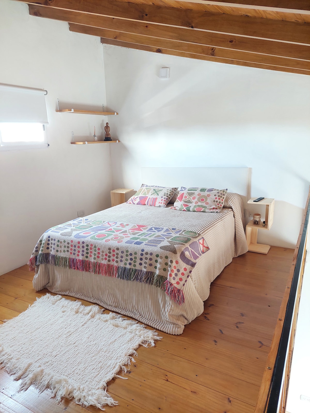 A bright bedroom features a spacious bed adorned with a decorative blanket and patterned pillows. Shelves are mounted on the wall, and a soft area rug lies on the wooden floor. Natural light enters through a window, highlighting the clean and simple design.