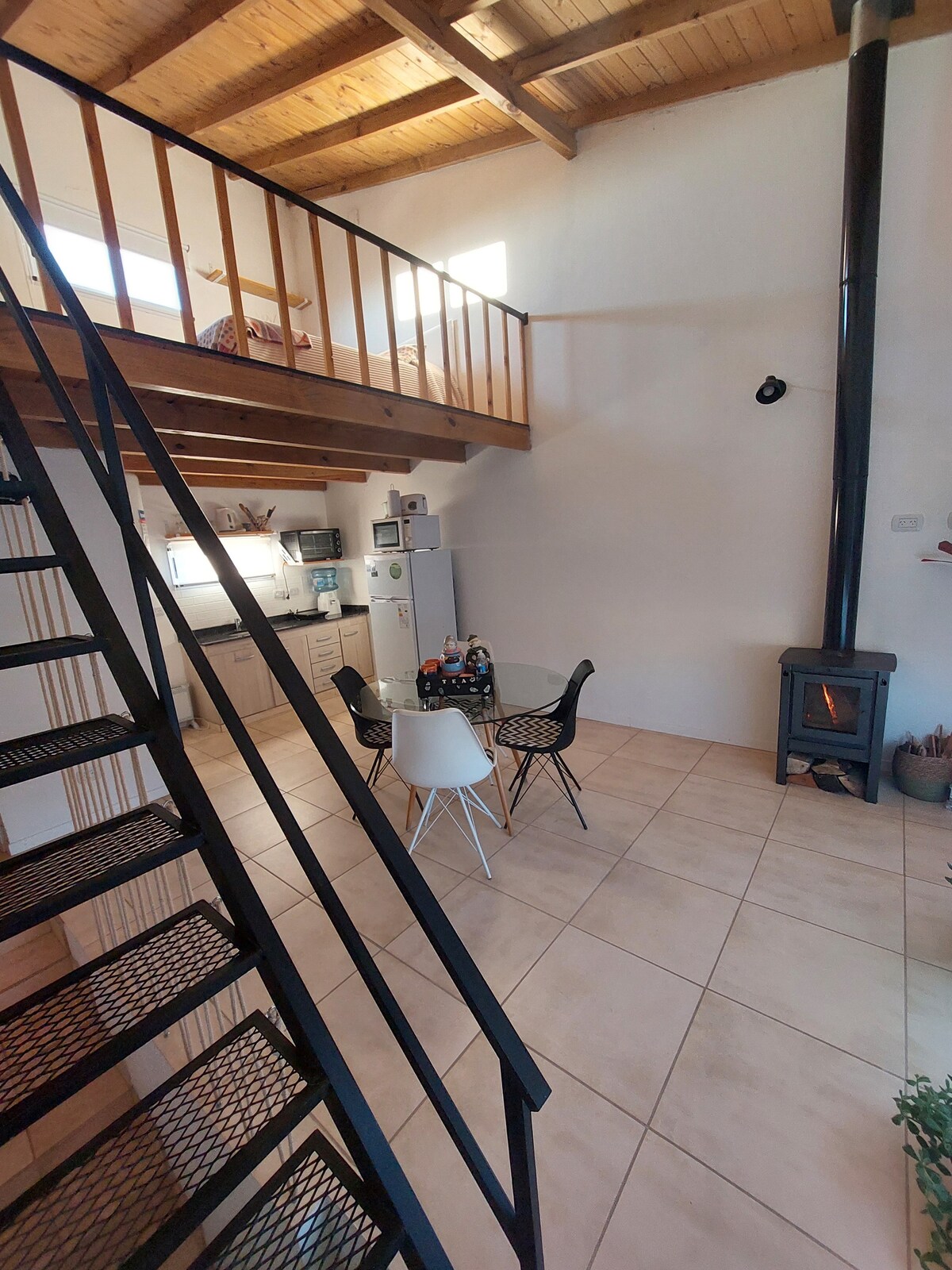 A bright, open living space features a staircase leading to a lofted sleeping area. A circular dining table with four chairs is positioned near a compact kitchen. A wood-burning stove provides warmth, accentuating the natural wood beams overhead.