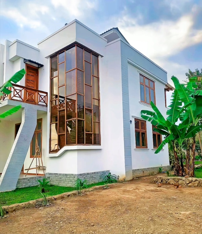 The modern villa exterior features large windows that allow natural light to fill the interior. Lush green banana plants surround the property, enhancing the serene outdoor setting. A driveway leads to the entrance, showcasing the villa's spacious and inviting design.