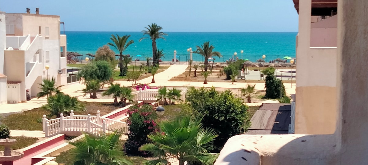 A view from the terrace showcases the vibrant blue sea in the distance, with palm trees and landscaped grounds framing the scene. Pathways meander through the greenery, leading towards the beach, while colorful umbrellas are visible along the shoreline.