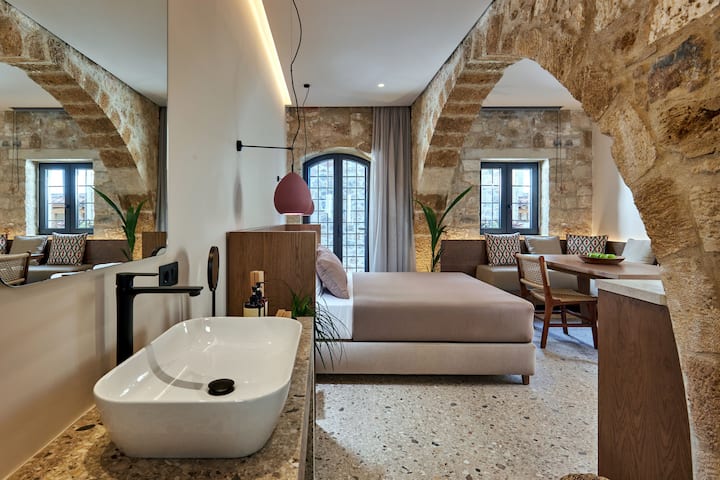 フロントゲストハウスの寝室。 素朴な石壁と洗練されたモダンなデザイン要素を融合させたエレガントな就寝エリアは、建物の3つの側面から流れる豊富な自然光に囲まれています。