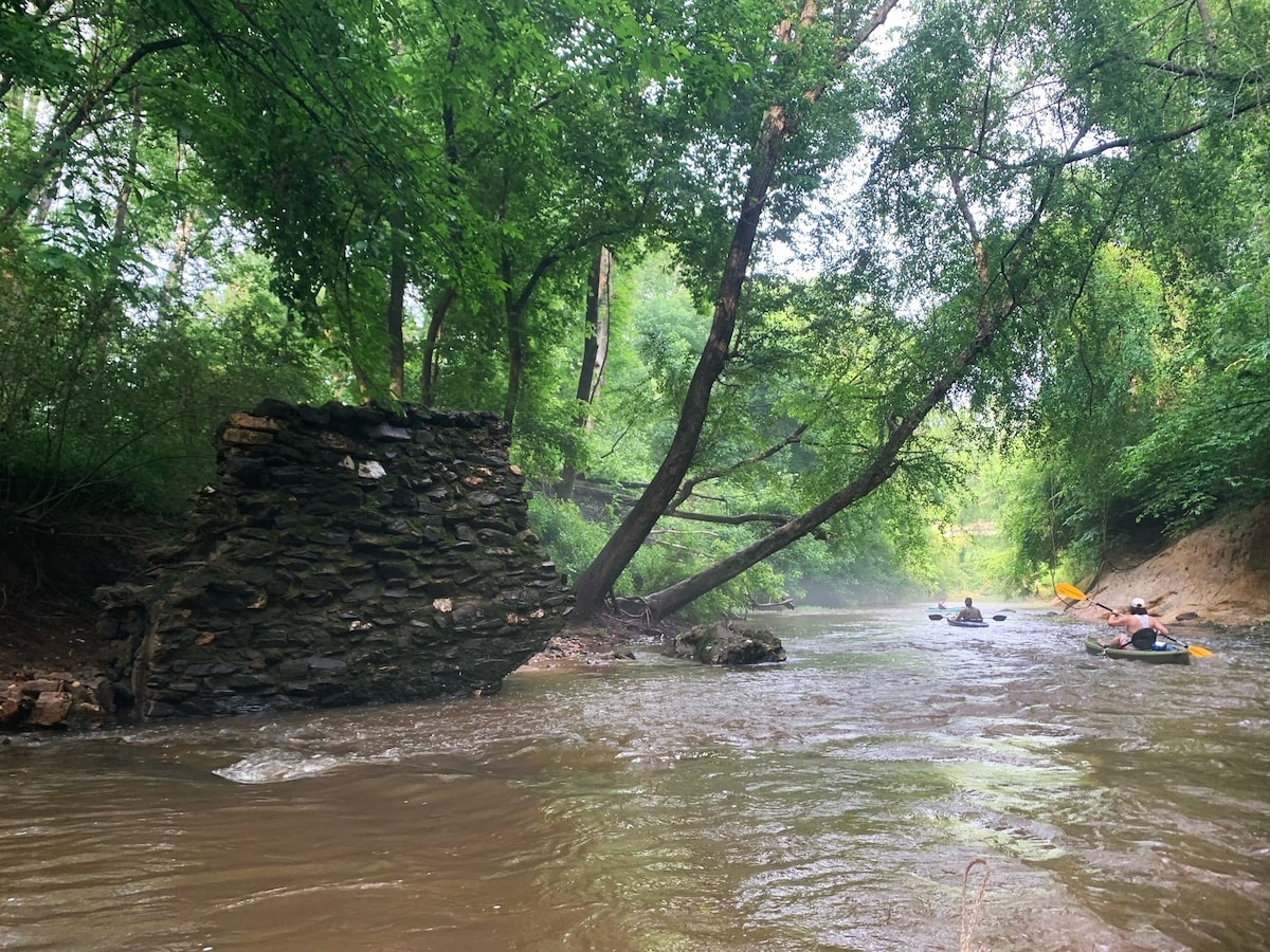 The image captures a winding creek framed by lush greenery, featuring a stone structure partially submerged along the bank. Canoers can be seen navigating the gentle waters, surrounded by an abundance of trees and foliage, creating a peaceful natural environment.