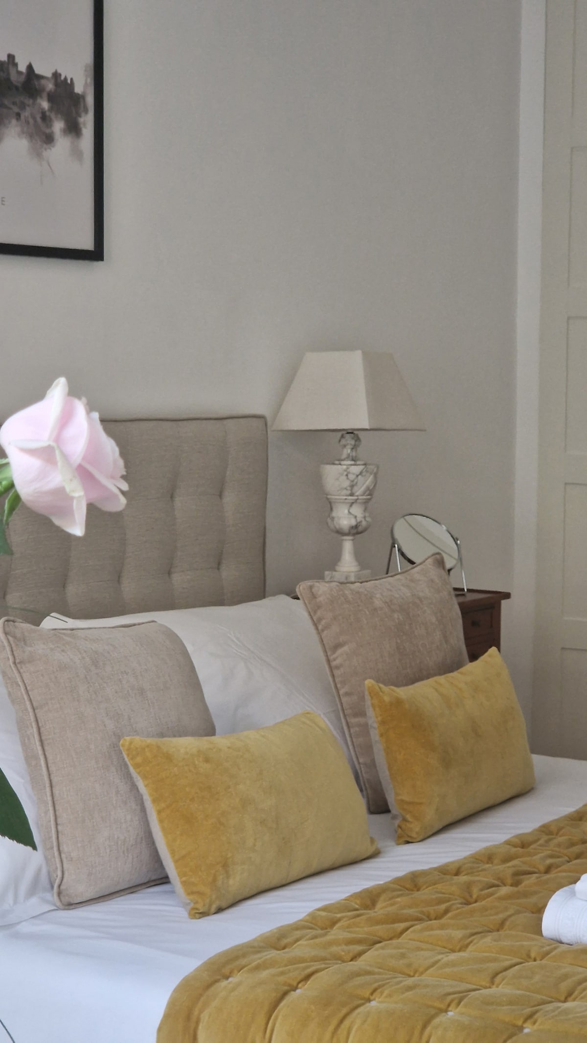 A elegantly furnished sleeping area is presented, featuring a spacious bed with soft, neutral bedding and decorative pillows in warm hues. A bedside lamp and a small mirror add functionality, while a delicate flower in the foreground introduces a touch of natural beauty.
