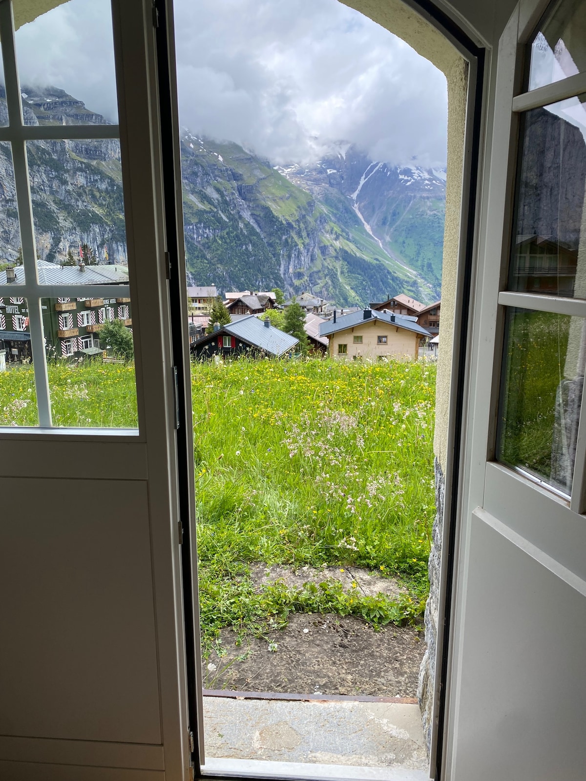 An open door frames a lush green landscape, showcasing rolling hills and the imposing mountains in the distance. Clouds gather above the peaks, and a colorful array of houses can be seen nestled in the valley below.