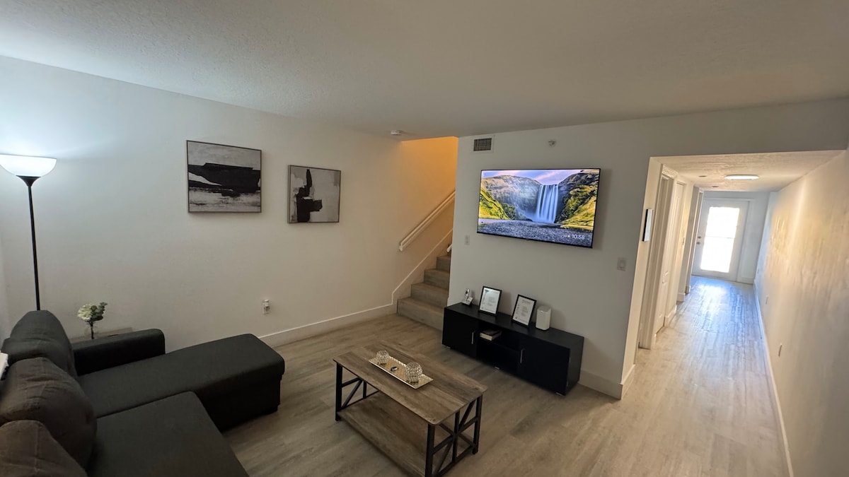 An inviting living area features a sectional sofa and a modern coffee table positioned on a light-colored floor. A wall-mounted television displays a scenic landscape, while simple artwork adorns the walls. Stairs lead to the upper level, enhancing the open layout.