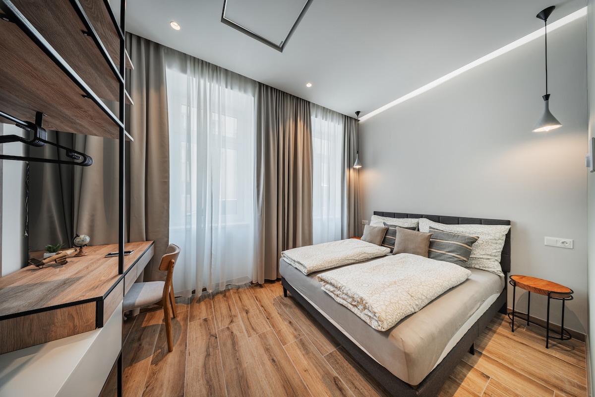 A modern studio features a spacious bed with neutral-coloured bedding and decorative pillows. Natural light filters through sheer curtains, illuminating the wood flooring. A wooden desk and chair are positioned near the window, complemented by a small side table.