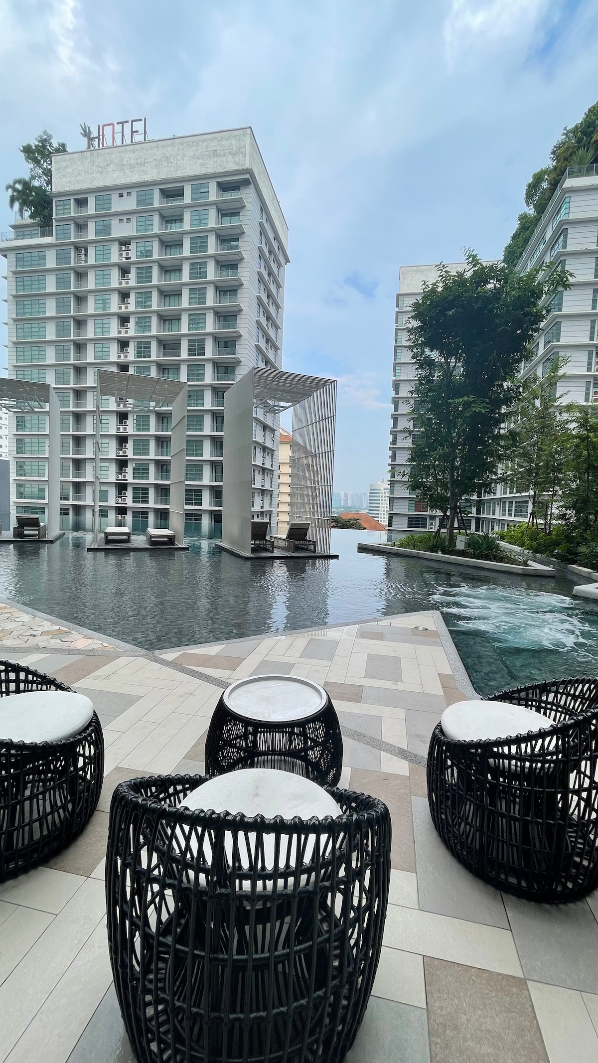 An outdoor area features modern, black wicker seating arranged around small round tables. The setting overlooks a tranquil pool, surrounded by lush greenery and contemporary buildings. Water softly reflects the blue sky, creating a serene atmosphere.
