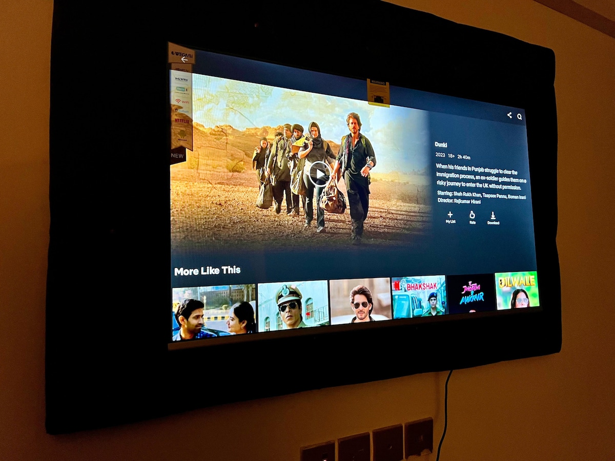 A Smart LED television is mounted on the wall, displaying a variety of movie options. The screen features a group of people in a landscape, with videos available below for selection. Multiple remote controls are placed neatly on a nearby console.