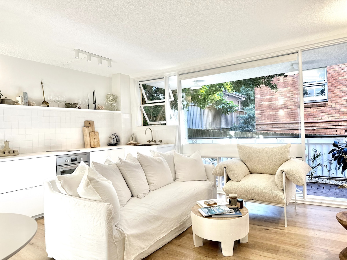 A bright living area is featured, showcasing a comfortable white sofa adorned with plush cushions. A cozy armchair complements the seating arrangement. Large glass doors create an open connection to the outside space, allowing natural light to fill the room.
