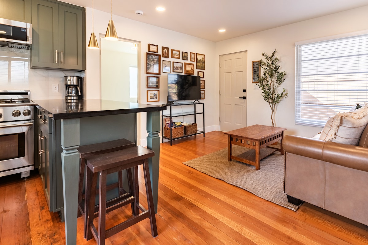 An open living space features a modern kitchen with dark cabinetry and stainless steel appliances. The wooden dining table is paired with two stools. A cozy seating area includes a brown leather sofa and a coffee table, set against a gallery wall of framed photos.
