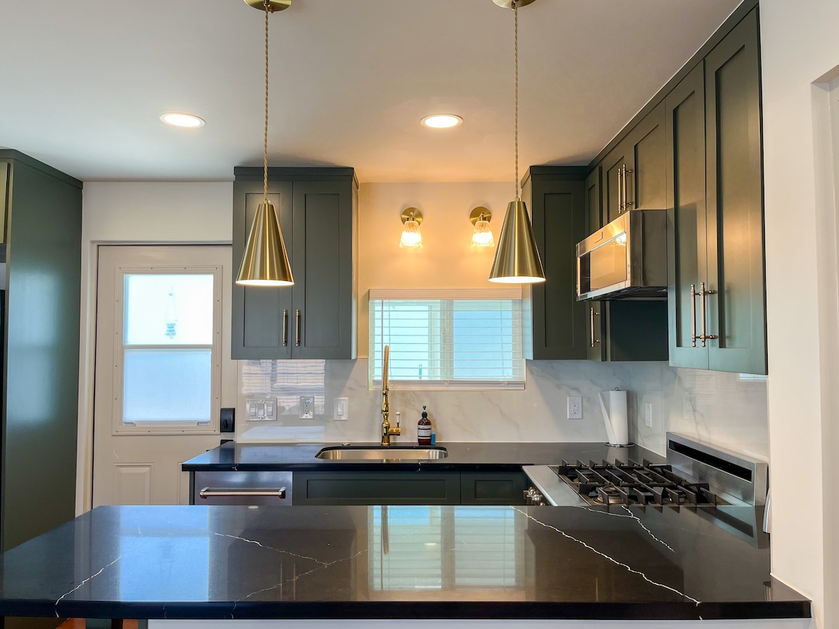 The kitchen features modern cabinetry with dark green finishes and a spacious black countertop. Pendant lights hang from the ceiling, creating a bright atmosphere. A window above the sink provides natural light, and a gas stove is installed beneath a stainless steel hood.