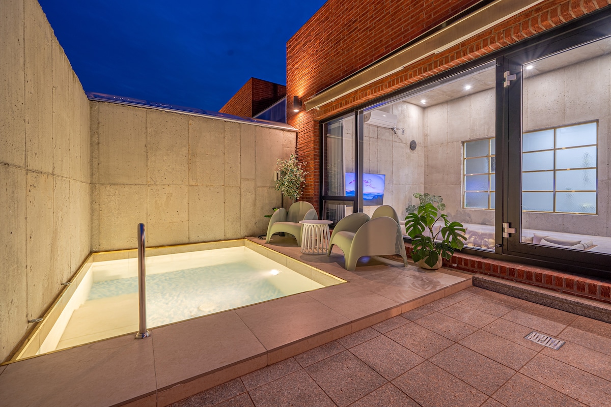 An outdoor area features a small private pool, surrounded by modern seating arrangements. The space is illuminated under a twilight sky, with a textured concrete wall in the background. Large glass doors provide views into the interior living area.