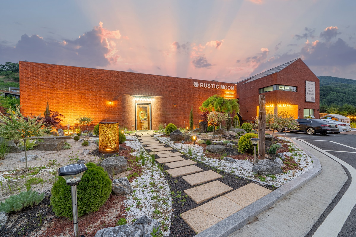 The exterior of the Rustic Moon residence is captured during sunset, showcasing a brick building surrounded by landscaped gardens. A stone pathway leads to the entrance, with decorative plants and stones framing the area, while soft rays of light create a serene ambiance.