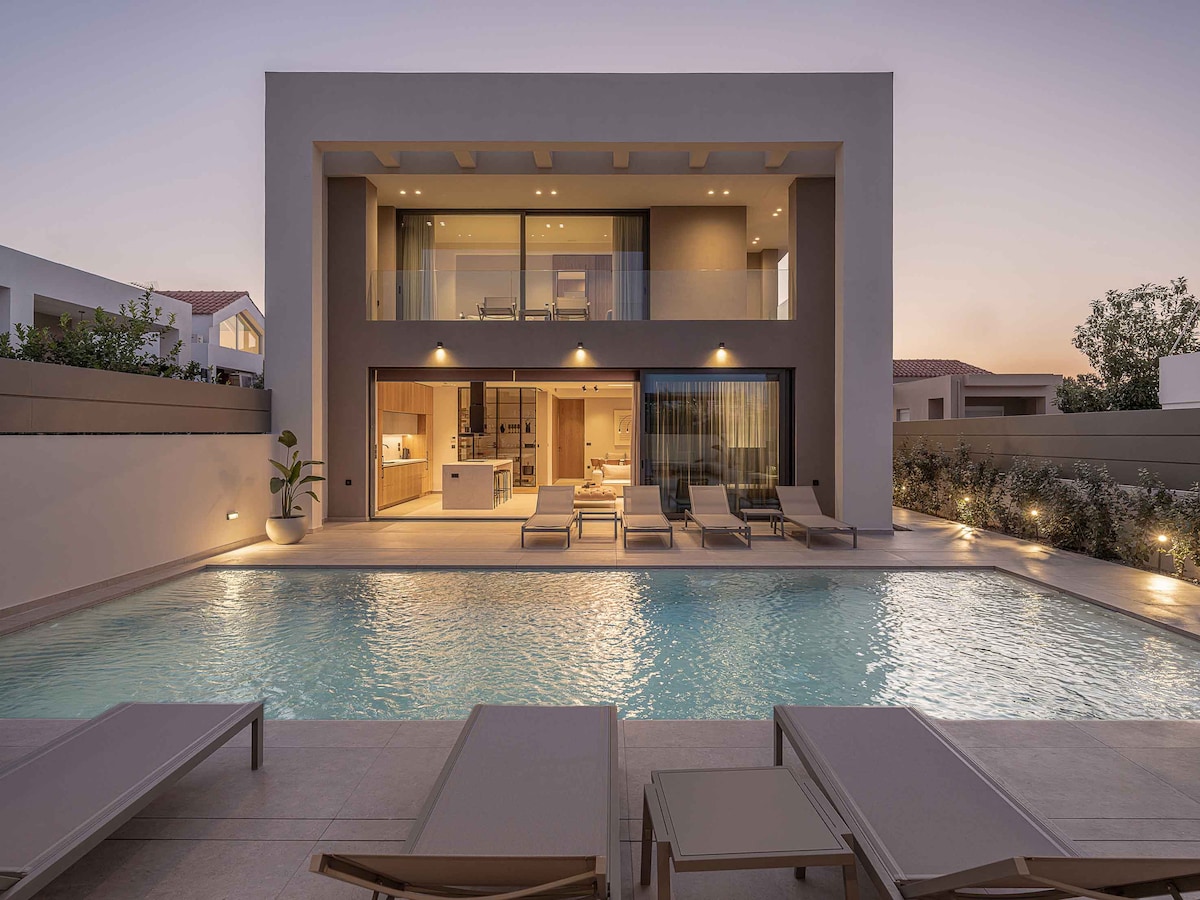 The villa features a modern architectural design, with large glass doors providing open views of the outdoor area and pool. Sun loungers are positioned around the heated swimming pool, which is illuminated in the evening light, enhancing a serene ambiance.