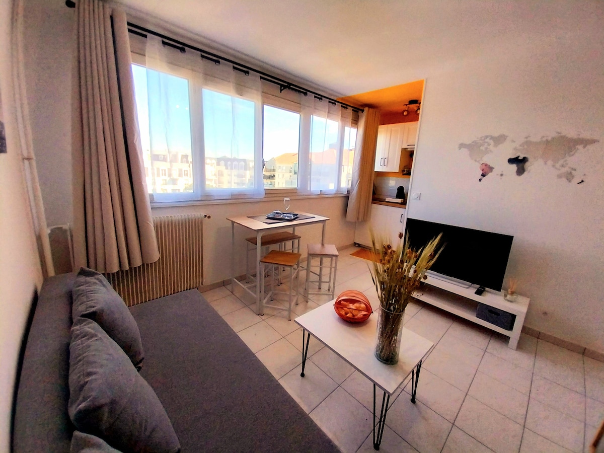 A sunlit living area features a comfortable gray sofa and a small dining table with two stools. A television sits on a minimalist white coffee table, and a decorative vase with dry plants adds a touch of warmth. Large windows fill the space with natural light.