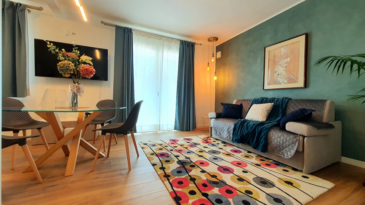 A modern living space features a comfortable sofa adorned with decorative pillows, complemented by a colorful area rug. A glass dining table with stylish chairs occupies the room, while large windows allow natural light to illuminate the space.